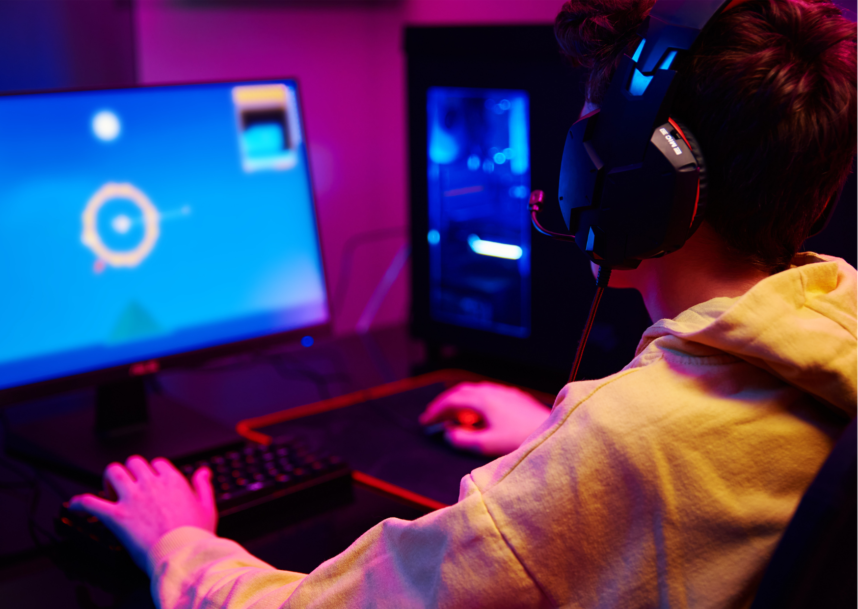 back of a person in a yellow hoodie waering gaming headphones with screen and gaming at a pc